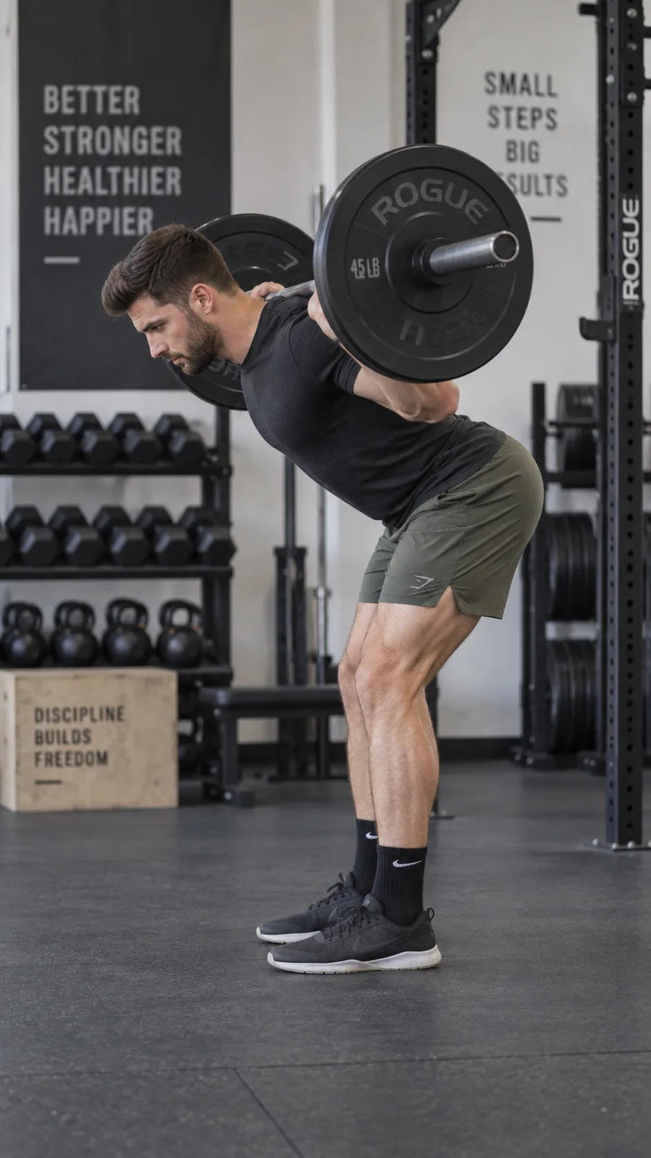 A male athlete performing a good morning exercise with a barbell across his upper back in a gym setting, demonstrating proper hip hinge form with a neutral spine and soft knees, side-on view, natural gym lighting