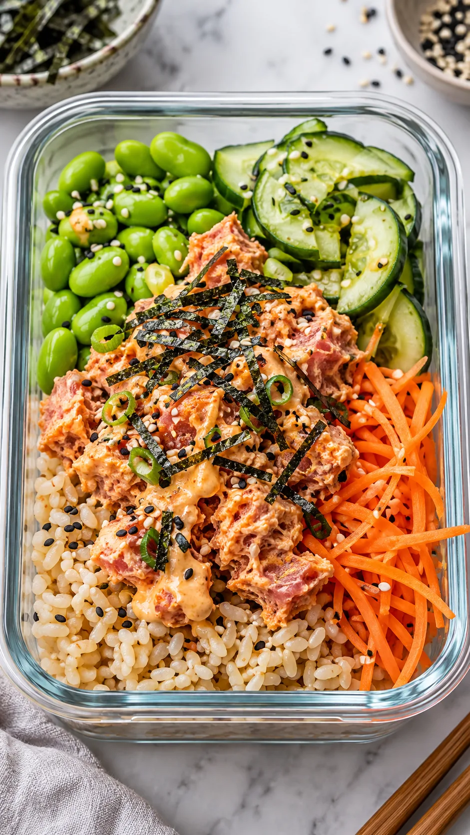 A styled food photograph of a deconstructed spicy tuna sushi bowl packed in a meal prep dish, showing flaked spicy tuna, brown sushi rice, edamame, sliced cucumber, shredded carrots, and nori strips