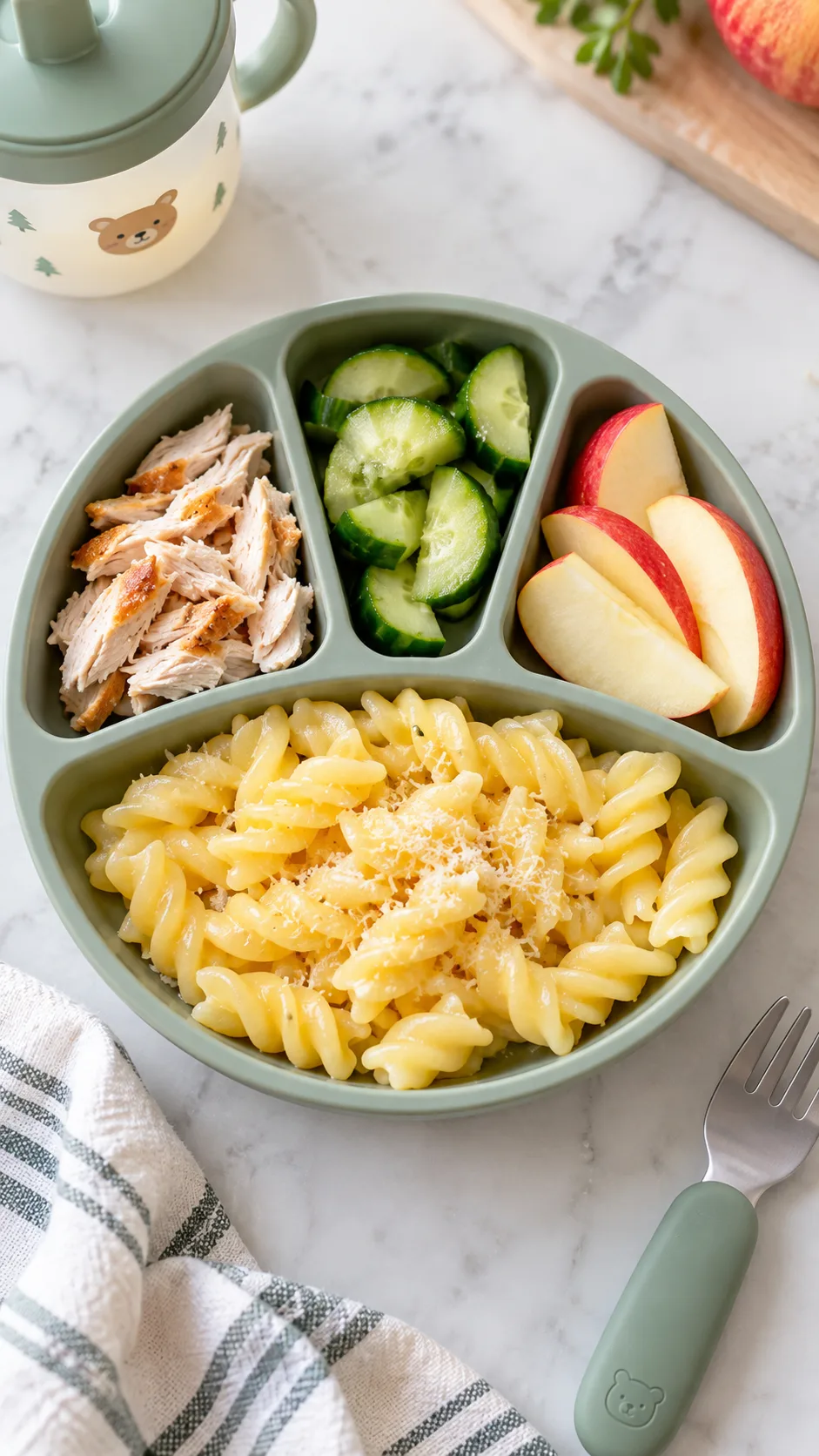 The Toddler Meal Plan That Finally Gets Picky Eaters to Eat 3 Overhead food photography of a toddler divided plate showing a picky eater meal formula: familiar buttered pasta, small portion of shredded chicken, cucumber half-moons, and apple slices; simple labels implied through arrangement, bright clean daylight, realistic family meal